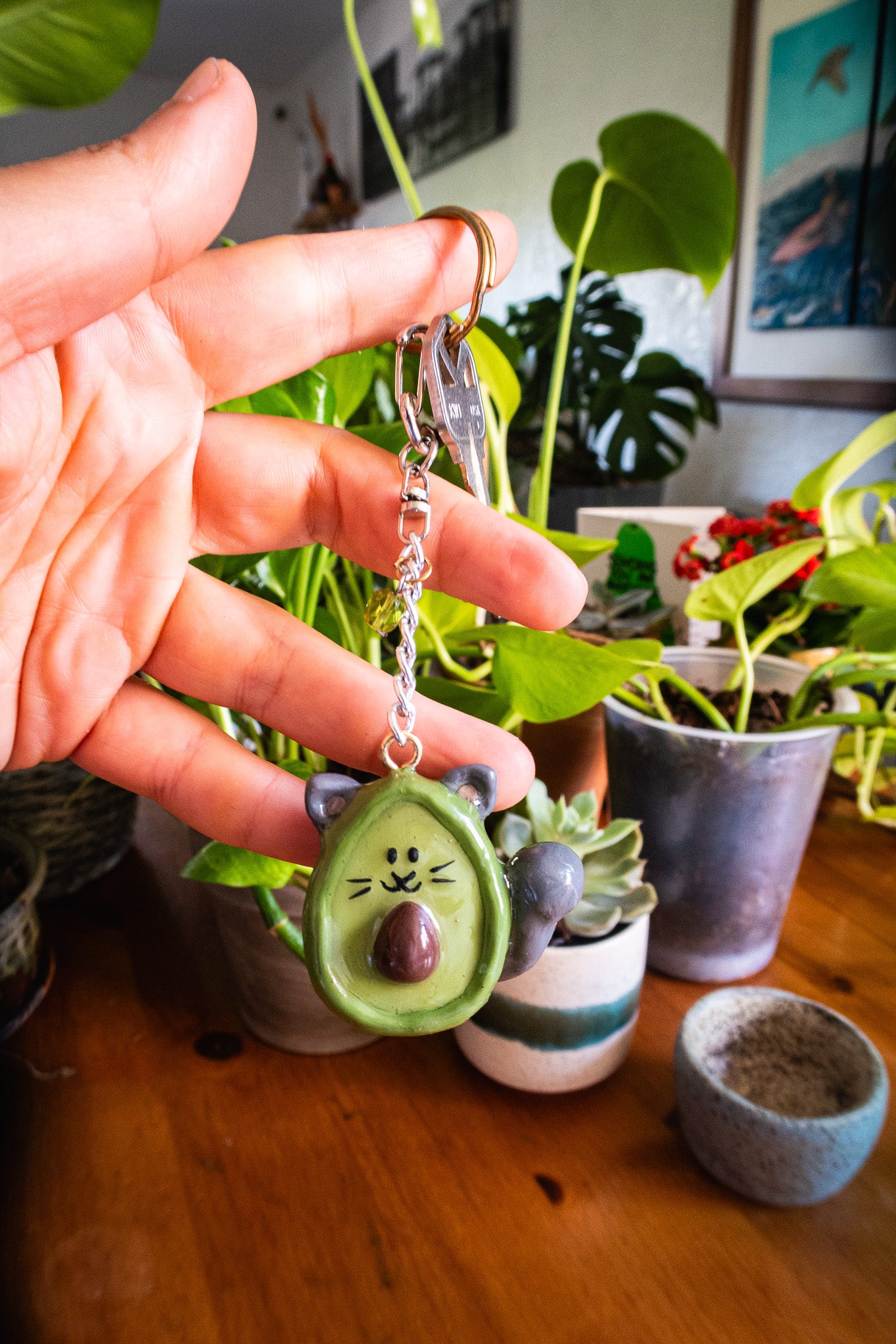 Hand holding a small green cat-avocado shaped keychain surrounded by potted plants.