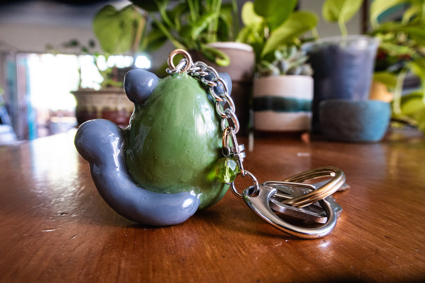 Back of the polymer clay cat keychain shaped like avocado on a wooden surface with plants in the background