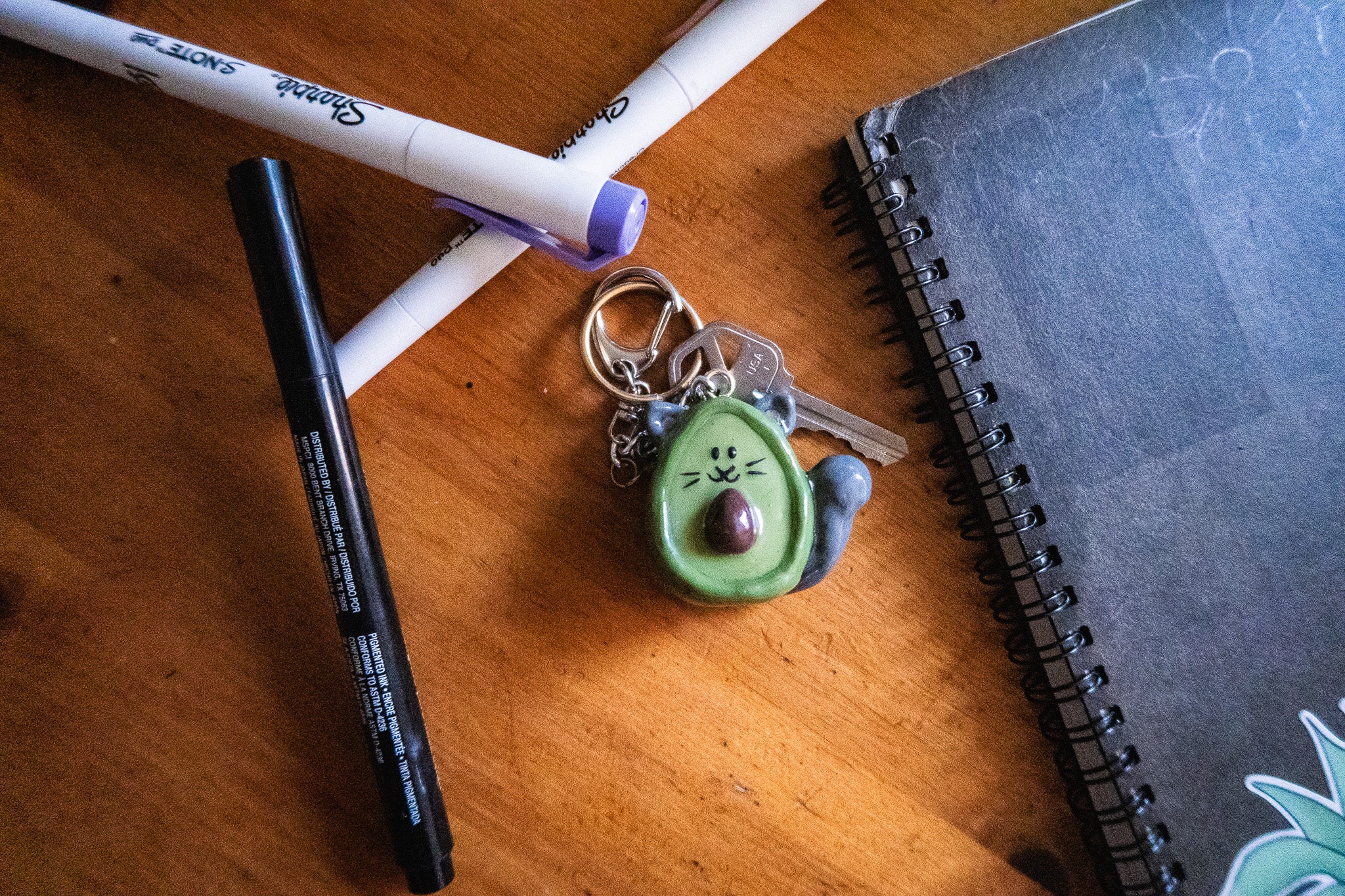 cat-avocado-shaped keychain on a wooden surface with markers and a notebook.