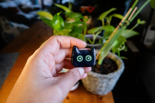 Hand holding a small black cat-shaped polymer clay magnet with green eyes in front of potted plants.