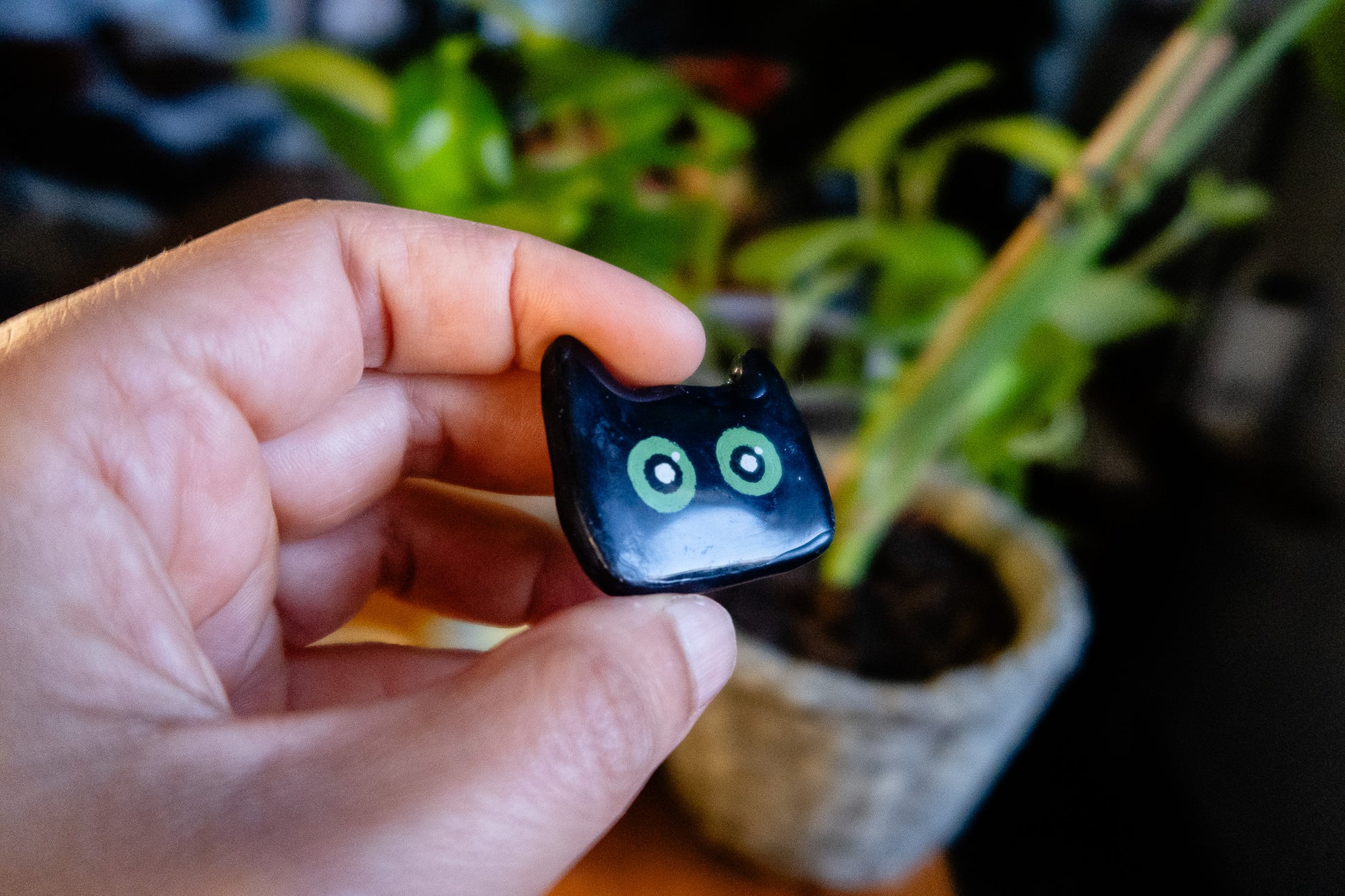 Hand holding a small black cat-shaped polymer clay magnet with green eyes  against a blurred indoor background
