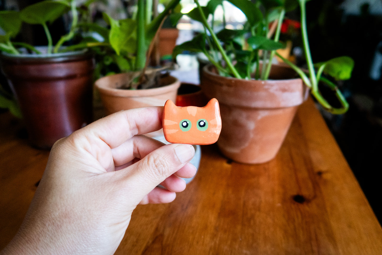Hand holding a small orange cat-shaped polymer clay magnet with potted plants in the background