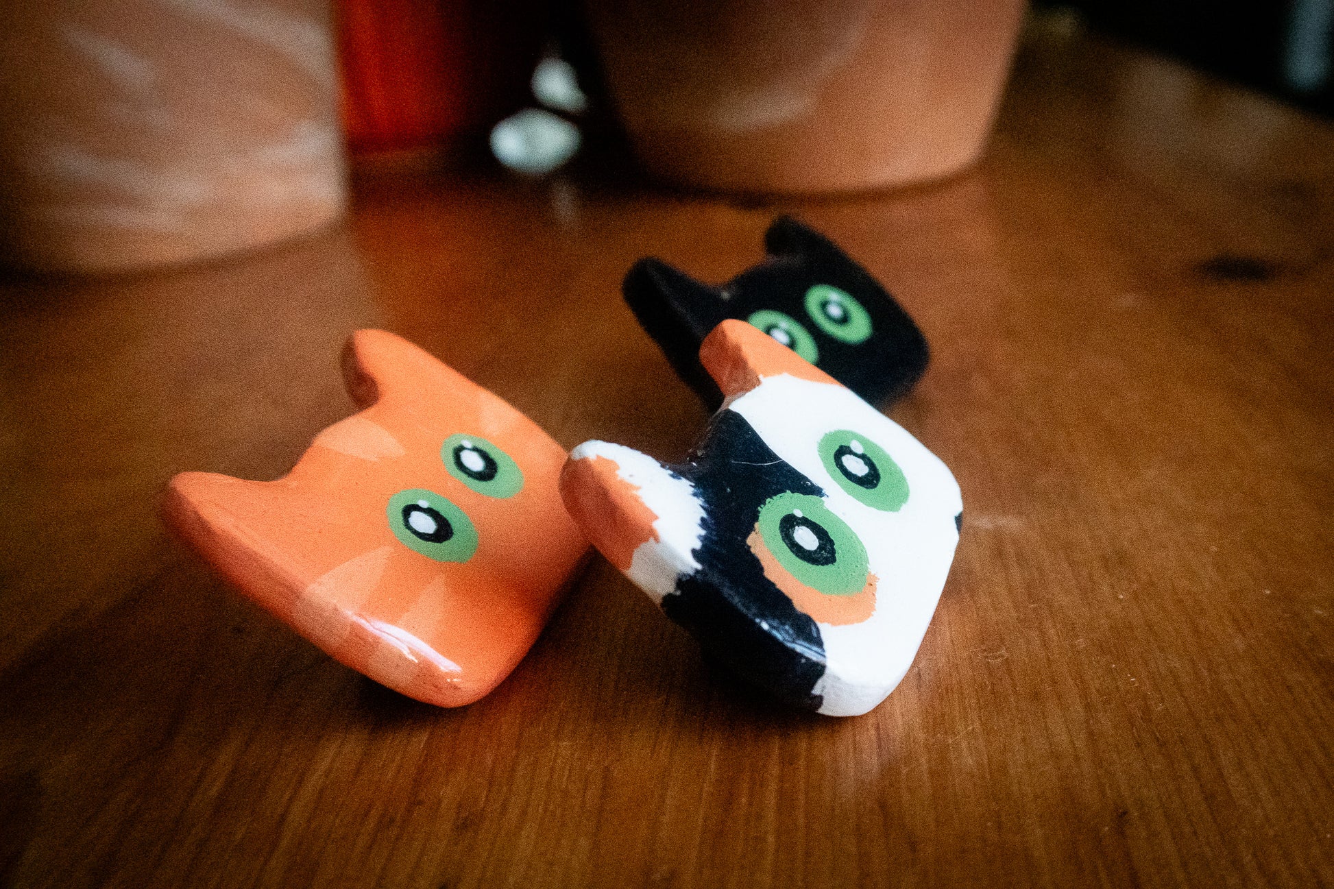 Three small cat figurines, one orange , one black and one black and white, on a wooden surface.