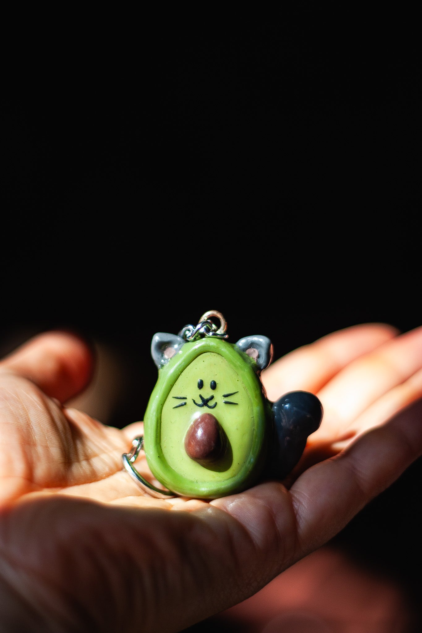 Avocado-shaped keychain with cat ears held in a hand against a dark background